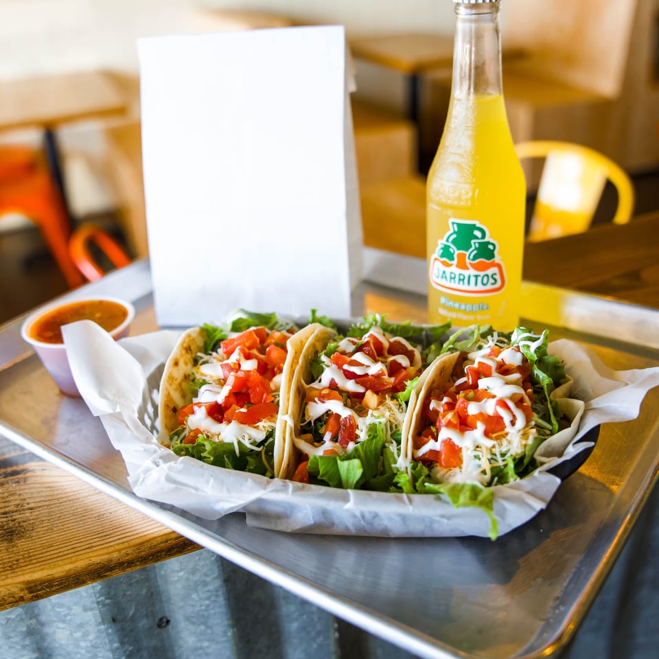 Three soft tacos topped with lettuce, cheese, diced tomatoes, and sour cream sit beside a Jarritos pineapple soda on a restaurant table.