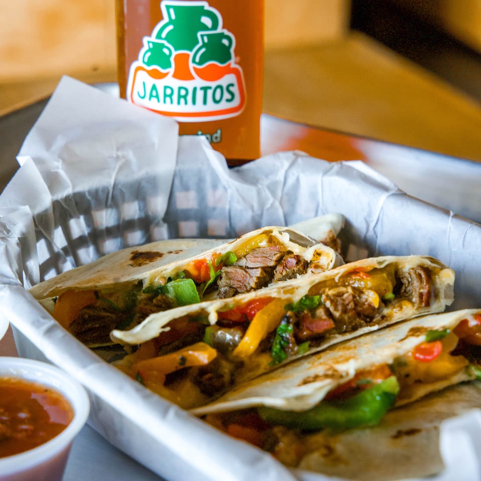 Grilled beef tacos with peppers and onions served in foil alongside a Jarritos orange soda and dipping sauce.