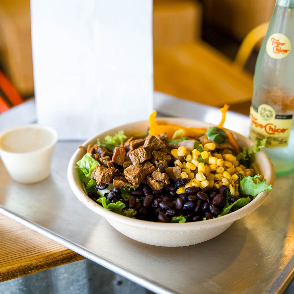 Bowl of leafy greens topped with grilled meat, black beans, corn, and vegetables beside a bottled drink.