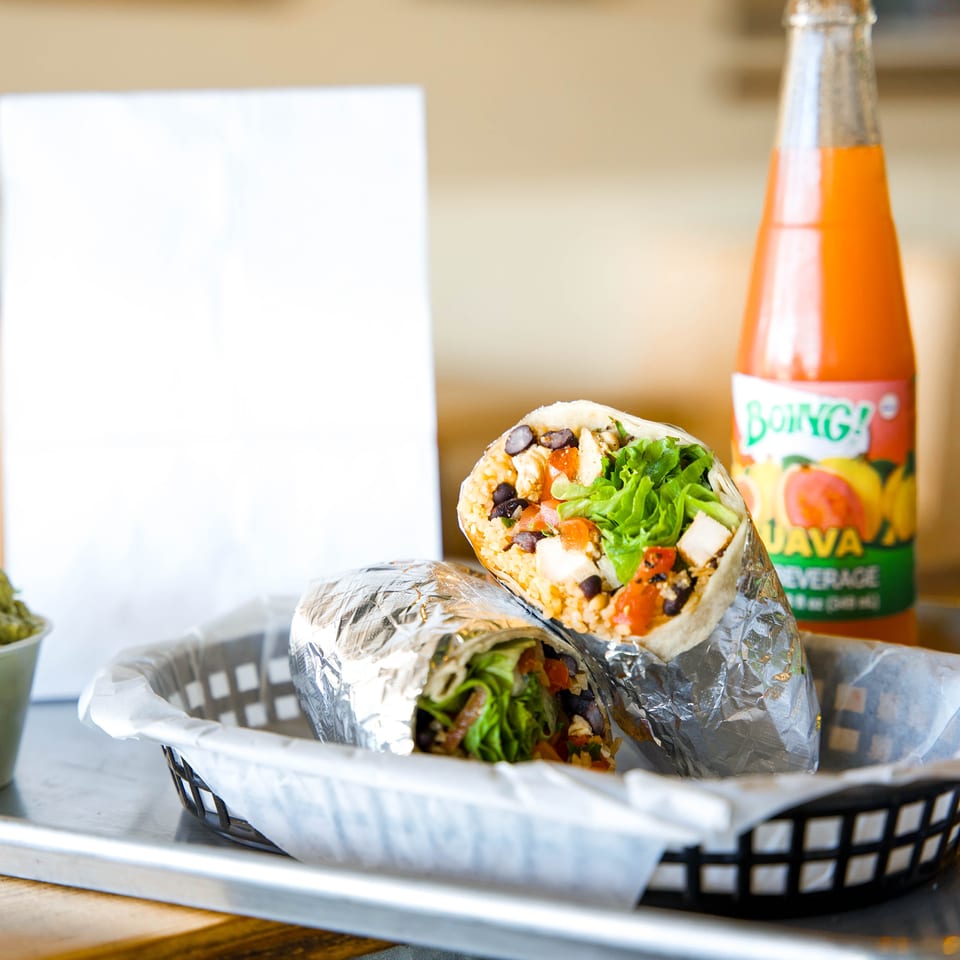 Foil-wrapped burritos filled with rice, black beans, vegetables, and lettuce sit in a basket beside a bottled orange soda.