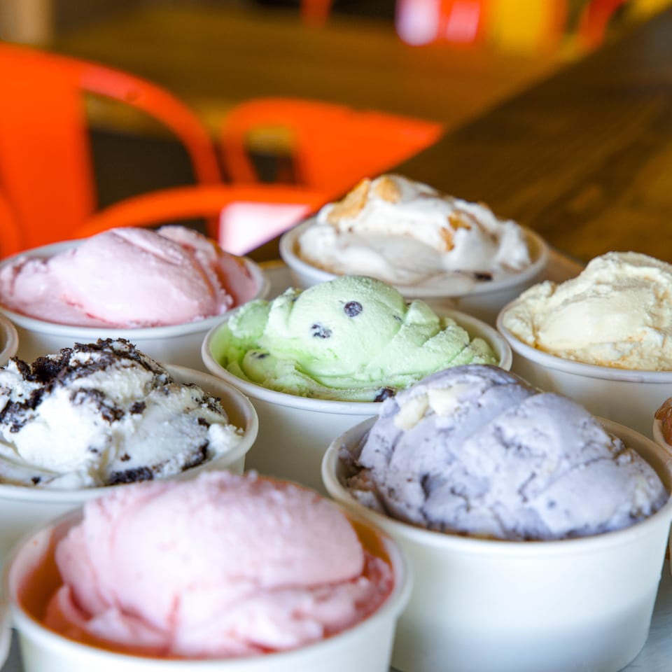 Colorful scoops of ice cream in white bowls displayed on a counter against a softly blurred restaurant interior.